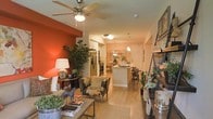 This single-floor residence is designed around an open-concept layout that connects the living area, dining space, and kitchen in one continuous, flowing arrangement. The living room draws immediate attention with a striking terracotta-red accent wall, which adds warmth and visual depth to the space. A ceiling fan with a built-in light fixture is centrally positioned overhead, contributing both functionality and a finishing architectural touch.

Light-toned hardwood flooring runs throughout, unifying each zone and enhancing the sense of spaciousness. The living area is furnished with patterned accent chairs and a coffee table, complemented by lush indoor greenery that adds a natural, lively element. A side table with a table lamp provides ambient lighting alongside the natural tones of the room.

The kitchen, visible from the living area, features a white island with bar seating, stainless steel appliances including a refrigerator and range, and white cabinetry. Pendant lighting hangs above the island, creating a defined dining and prep zone. The open sightlines from front to back make the space feel bright and connected throughout.