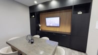 This refined single-floor space showcases a contemporary design aesthetic defined by clean lines, premium materials, and a carefully curated palette of dark and warm tones. The centerpiece of the space is a striking feature wall that combines rich wood-grain paneling with matte black cabinetry, housing a wall-mounted flat-screen display — ideal for presentations or collaborative viewing. Flanking the feature wall, floor-to-ceiling built-in cabinetry with sleek hardware provides ample storage while maintaining a streamlined, professional appearance. Open shelving niches offer space for decorative accents, adding subtle warmth to the room. A large marble-topped conference table takes center stage, surrounded by white upholstered seating that provides an elegant contrast to the darker surrounding elements. Recessed ceiling lighting ensures the space is well-illuminated, casting a clean, focused light throughout. The overall ambiance is polished and professional, balancing functionality with sophisticated interior design. Every detail — from the material choices to the furniture selection — reflects a deliberate effort to create an environment that is both highly practical and visually impressive.