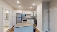 Located at 250 Pine Knot Ln, Unit 305, in Graham, NC, this single-floor residential unit offers a thoughtfully designed layout with modern finishes throughout. The kitchen serves as a central highlight, featuring crisp white shaker-style cabinetry, granite countertops, and a full suite of stainless steel appliances including a refrigerator, electric range with a sleek overhead hood, built-in microwave, and dishwasher. A granite-topped island provides additional workspace and storage, while rich wood-look flooring anchors the space with warmth and durability. The open layout connects the kitchen seamlessly to a spacious living area finished with plush neutral carpeting, creating a comfortable and versatile space. The bedroom is generously sized, carpeted for comfort, and includes a ceiling fan, natural light from window blinds, and access to an expansive walk-in closet outfitted with wire shelving systems offering ample storage. The bathroom is well-appointed with tile flooring, a tub-shower combination with white tile surround, granite vanity, and built-in wire shelving for added convenience. Recessed lighting throughout contributes to a clean, modern ambiance across the entire unit.