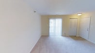 This single-floor space presents a clean, functional layout with a light and airy ambiance throughout. The main living area is finished with soft wall-to-wall carpeting in a neutral tone, complementing the warm, cream-colored walls that give the space a welcoming feel. A large sliding glass door serves as a focal point, fitted with vertical blinds that allow for adjustable natural light while providing a direct connection to the outdoors. The room benefits from generous natural light that brightens the interior and enhances the open, uncluttered atmosphere. A wall-mounted light fixture adds a subtle decorative touch, while the smooth white ceiling contributes to the sense of openness. A closet with louvered doors is conveniently situated along one wall, offering practical built-in storage. The overall design is straightforward and versatile, providing a neutral canvas well-suited for a variety of furnishing arrangements. The combination of soft flooring, natural light, and sliding outdoor access makes this a comfortable and adaptable living space.