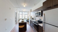 This single-floor unit showcases a thoughtfully designed open-concept layout with distinct industrial-modern character. Exposed ductwork and ceiling infrastructure lend a loft-inspired aesthetic throughout, complemented by light hardwood flooring and crisp white walls.

The kitchen is well-appointed with dark espresso cabinetry, white countertops, a subway tile backsplash, and a full suite of stainless steel appliances including a refrigerator, electric range, microwave, and dishwasher. A central island with bar seating provides additional prep space and casual dining. In-unit stacked washer and dryer units are conveniently accessible nearby.

The living area is anchored by a large picture window that frames sweeping urban skyline views and fills the space with abundant natural light. The bedroom continues this theme with oversized windows offering city vistas, creating a bright and airy atmosphere. A second, more intimate bedroom provides a quieter retreat with its own window access.

The bathroom features clean, minimalist finishes including large-format floor tiles, a floating dark-wood vanity with a marble-style countertop, a large mirror, and open white shelving for organized storage. The overall ambiance is contemporary, light-filled, and urban.