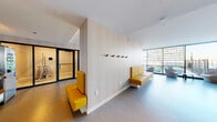 Located at 1200 S Indiana Ave in Chicago's South Loop, this single-floor amenity space is designed to serve both residents and their pets. The layout is divided into distinct functional zones, each thoughtfully appointed with contemporary furnishings and cohesive design elements.

The lounge area features patterned, warm-toned wallcovering that runs throughout the space, providing visual continuity. Bold yellow upholstered seating—including banquettes and modular ottomans—anchors one section of the lounge, complemented by wooden wall-mounted coat hooks. Floor-to-ceiling windows along one wall flood the space with natural light and offer open views of the surrounding Chicago skyline and an adjoining outdoor terrace furnished with additional seating.

A separate lounge zone transitions into a cooler color palette, featuring a blue sectional sofa, rounded gray ottomans, and low oval coffee tables atop blue-toned flooring—creating a calm, relaxed atmosphere. Soft recessed lighting and wall sconces provide warm illumination throughout.

A dedicated pet care area includes a stainless steel Evolution Dog Wash station, wood cabinetry with a dark countertop, open shelving with green pipe accents, and potted plants. An enclosed glass-paneled room, visible from the main corridor, provides additional private space. Restroom access is also available within the suite.