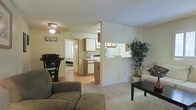 This single-floor residence offers a practical and welcoming layout centered around an open-concept living area. The carpeted living room provides a cozy gathering space, furnished with plush seating and a coffee table, and is brightened by a ceiling fan with integrated lighting. A large decorative plant adds a natural accent to the corner of the room.

Adjacent to the living area, a dining space accommodates a dark-toned table and chairs, seamlessly connecting the social zones of the home. The kitchen is positioned just beyond a partial dividing wall, featuring warm wood-toned cabinetry, white appliances, and a pass-through counter that maintains visual openness between the kitchen and the main living space. The kitchen's hard-surface flooring contrasts neatly with the carpet in the adjoining areas.

A hallway visible from the main living area leads to the private quarters of the home, suggesting a well-organized floor plan that separates the living and sleeping spaces. Throughout, the interior is finished in soft, neutral tones that create a bright and airy ambiance. The textured ceiling, clean white walls, and consistent trim details contribute to a cohesive and functional residential environment.