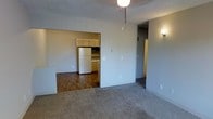 This single-floor residential space offers a practical and straightforward layout designed for comfortable everyday living. The main living area is finished with wall-to-wall carpet in a neutral tone, creating a warm and understated atmosphere. A textured ceiling and a central ceiling light fixture add to the functional character of the space, while multiple electrical outlets are conveniently distributed throughout.

An open rectangular passageway connects the living area directly to the kitchen, which features wood-tone cabinetry along both the upper and lower sections, providing ample storage. The kitchen is equipped with a full-size white refrigerator and includes countertop workspace, with flooring transitioning to hardwood-style planks that contrast with the carpeted living space.

A hallway, visible from the living area and accented by a wall-mounted light fixture, leads toward additional areas of the unit, suggesting a separation between the common living spaces and more private rooms. The neutral gray wall tones throughout create a clean, cohesive look that offers a versatile backdrop for a variety of interior styles. Overall, the space presents a functional, no-frills design well-suited for practical residential use.