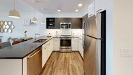 This well-designed one-bedroom, one-bathroom unit showcases a modern aesthetic throughout. The open-concept kitchen is a standout feature, equipped with stainless steel appliances including a full-size refrigerator, microwave, and range. Dark upper cabinetry contrasts elegantly with crisp white lower cabinets, while a spacious island with dark countertops and pendant lighting provides seating for three. Wood-look flooring and a white subway tile backsplash complete the kitchen's contemporary look.

The living area benefits from large windows that invite abundant natural light and offer views of the surrounding urban landscape. Dark hardwood-style flooring flows throughout, grounding the bright, white walls. The bedroom continues this theme, featuring large windows and direct access to a walk-in closet with built-in shelving and hanging rods for practical storage.

The bathroom presents a clean, minimalist design with large-format tile flooring, a white vanity with ample drawer storage, and a combined tub and shower. Throughout the unit, the neutral palette and modern finishes create a cohesive, contemporary living environment well-suited for urban living.