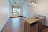 Located at 324 Clifford St in the heart of Detroit, this single-floor unit showcases an industrial-modern aesthetic that blends raw architectural character with contemporary finishes. The open living area is anchored by rich hardwood flooring that runs throughout, while exposed ceiling beams painted white add a loft-like quality and a sense of vertical openness to the space.

The kitchen is a functional centerpiece, featuring a peninsula island with a quartz countertop, an undermount stainless steel sink, and a dishwasher integrated beneath dark cabinetry. Stainless steel appliances—including a range, over-the-range microwave, and refrigerator—are paired with white upper cabinets and light stone countertops. A multi-bulb brass pendant light fixture overhead adds a warm, industrial-chic touch.

The main living area benefits from natural light streaming through large windows framed in black, offering views of the surrounding Detroit skyline. A wall-mounted mini-split unit provides climate control.

The bathroom is cleanly finished with gray accent walls, a walk-in shower with built-in shelving, a dark wood vanity with a white countertop, and neutral tile flooring, creating a practical and polished space.