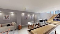 Located at 45 Main St in Yonkers, NY, this versatile lounge and multi-use room is a thoughtfully designed amenity space that seamlessly blends social, recreational, and entertainment functions. The single-floor layout is anchored by soaring double-height ceilings and large-format porcelain tile flooring throughout, giving the space an open, airy feel.

A standout feature is the dramatic natural wood-clad accent wall, which adds warmth and texture against crisp white walls. A tiered wooden bleacher-style seating area with cushioned seats serves as a screening or presentation zone, positioned alongside a large wall-mounted display. Opposite, a generously sized communal bar counter with a rich wood surface is surrounded by modern bar stools and topped with colorful pendant lighting in muted blue and yellow tones. A kitchenette with a sink and counter space supports food and beverage service.

The lounge area features a plush sectional sofa, layered rugs, a marble coffee table, and playful pop-art wall décor, creating a relaxed, social atmosphere. A foosball table and shuffleboard court add recreational options. Recessed and accent lighting throughout ensures a warm, inviting ambiance, while the open floor plan allows flexible configurations for a variety of events and gatherings.