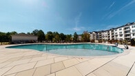 Located at 4101 Glenloch Creek in Matthews, NC, this thoughtfully designed outdoor amenity space offers a welcoming and well-appointed environment for relaxation and recreation. At the heart of the space is a generously sized resort-style swimming pool with a smooth stone tile surround, lined with lounge chairs perfect for sunbathing under the Carolina sky. The pool area is framed by mature trees and lush greenery, providing a natural backdrop that enhances the open-air atmosphere.

Adjacent to the pool, a spacious covered patio extends along the exterior of the building, supported by sturdy columns and finished with a clean concrete surface. This shaded outdoor area is furnished with wicker lounge chairs, cushioned seating, and wooden tables, creating an ideal setting for casual gatherings or quiet relaxation. Oversized decorative planters with vibrant florals add color and character throughout the space.

The patio also features an outdoor game area equipped with a large-scale stacking block game, encouraging social interaction among residents and guests. The building exterior showcases a modern design with white brick, board-and-batten siding, large picture windows with decorative teal curtains, and stone accents at the base. A gated entry with clear emergency access signage ensures safety throughout the amenity grounds.