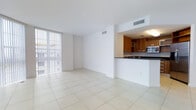 This single-floor space features an open-plan layout that connects the living area to the kitchen, creating a sense of spaciousness and flow. The interior is finished with large-format white ceramic tile flooring that extends throughout, reflecting light and enhancing the clean, modern aesthetic.

The living area is expansive and bathed in natural light from a floor-to-ceiling window fitted with vertical blinds, offering both brightness and privacy control. The crisp white walls and smooth ceiling, complete with recessed HVAC vents, contribute to the space's minimalist character.

The kitchen is defined by a raised breakfast bar with a dark countertop, providing a natural visual divider between the cooking and living zones. Warm-toned wood cabinetry lines the kitchen walls, offering ample storage, while the integrated microwave and built-in appliances add functional convenience. The contrast between the rich cabinetry and the white surroundings creates a focal point that anchors the space. Overall, the unit presents a bright, airy, and well-organized environment suited to modern living.