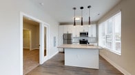 Located at 223 E Town St in Columbus, Ohio, this single-floor residence offers a thoughtfully designed open-concept layout with a clean, contemporary aesthetic. The kitchen serves as the heart of the space, showcasing crisp white cabinetry that extends to the ceiling for ample storage, paired with a full suite of stainless steel appliances including a refrigerator, range, and built-in microwave. A central kitchen island with a light-toned countertop provides additional workspace and casual seating potential, while three stylish pendant lights — featuring sleek, modern silhouettes — hang above, adding both function and visual interest. Natural light flows in through a window fitted with white blinds, brightening the space and complementing the neutral color palette. The flooring transitions from warm hardwood-style planks in the kitchen and common areas to plush carpeting in the adjacent rooms, offering a comfortable contrast. A glass-paneled interior door is visible leading to a neighboring room, adding an architectural detail that enhances openness while maintaining a sense of defined space. The overall ambiance is light, airy, and move-in ready, reflecting a modern urban lifestyle in the heart of Columbus.