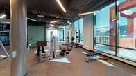 Located at 2 I St SE in Washington, DC, this contemporary fitness center offers a well-appointed, single-floor facility designed to support a full range of workout routines. The space is divided into two distinct areas: a strength and cardio equipment zone and a dedicated movement studio.

The equipment area features an extensive selection of Life Fitness machines, including cable systems, weight stacks, benches, and free weights. Rubber flooring provides durability and cushioning throughout, while an exposed ceiling with linear LED lighting lends an industrial character to the space. A wall-mounted flat-screen television adds a media element, and floor-to-ceiling windows along multiple walls flood the room with natural light and frame views of the surrounding Washington, DC skyline and neighboring developments.

The adjacent studio is a standout space, distinguished by a warm wood-slat ceiling, rich hardwood flooring, and walls of teal-painted accents. Full-length mirrors line one wall, making it well-suited for yoga, stretching, dance, or group fitness classes. Built-in low storage benches and yoga mat racks provide functional organization. Expansive glass windows wrap around the studio, offering sweeping outdoor views and abundant natural light that create an open, energizing atmosphere. Together, the two areas form a comprehensive, modern fitness facility with thoughtful design details throughout.