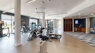 Located at 301 Silveron Blvd in Flower Mound, Texas, this fitness center offers a spacious, single-floor exercise facility thoughtfully designed to support a full range of workout routines. The open floor plan is divided into distinct functional zones, creating an organized and efficient layout for users of all fitness levels.

The strength training section features professional-grade Matrix equipment, including cable machines, weight benches, and free weight racks with barbell and plate storage. Full-length mirrors line the walls, enhancing the sense of space and allowing users to monitor their form. A dedicated functional training area is defined by a bold geometric-patterned area rug and includes a heavy punching bag, speed bag, wall-mounted storage for medicine balls and dumbbells, and a pull-up rig — ideal for high-intensity and combat-style workouts.

The cardio zone is well-stocked with treadmills, ellipticals, stair climbers, and additional Matrix cardio machines positioned to take advantage of the abundant natural light streaming through large floor-to-ceiling windows. These windows also provide direct access to an outdoor terrace with open views of the surrounding landscape. Ceiling fans are distributed throughout the space, promoting airflow and comfort during intense sessions. Light wood-tone flooring, clean white walls accented by dark feature walls, and warm wooden shelving add a modern, polished aesthetic to the overall environment.