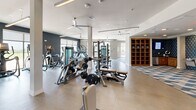 Located at 301 Silveron Blvd in Flower Mound, Texas, this fitness center offers a spacious, single-floor exercise facility thoughtfully designed to support a full range of workout routines. The open floor plan is divided into distinct functional zones, creating an organized and efficient layout for users of all fitness levels.

The strength training section features professional-grade Matrix equipment, including cable machines, weight benches, and free weight racks with barbell and plate storage. Full-length mirrors line the walls, enhancing the sense of space and allowing users to monitor their form. A dedicated functional training area is defined by a bold geometric-patterned area rug and includes a heavy punching bag, speed bag, wall-mounted storage for medicine balls and dumbbells, and a pull-up rig — ideal for high-intensity and combat-style workouts.

The cardio zone is well-stocked with treadmills, ellipticals, stair climbers, and additional Matrix cardio machines positioned to take advantage of the abundant natural light streaming through large floor-to-ceiling windows. These windows also provide direct access to an outdoor terrace with open views of the surrounding landscape. Ceiling fans are distributed throughout the space, promoting airflow and comfort during intense sessions. Light wood-tone flooring, clean white walls accented by dark feature walls, and warm wooden shelving add a modern, polished aesthetic to the overall environment.