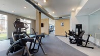Located at 210 S Main St in Kannapolis, North Carolina, this single-floor fitness center offers a comprehensive and thoughtfully designed workout facility. The space is organized into distinct functional zones that flow seamlessly into one another, defined by a curved flooring transition between light and darker gray rubber surfaces that adds a dynamic visual element to the layout.

The cardio zone is well-stocked with treadmills, stair climbers, and elliptical machines, positioned to take advantage of large windows that invite abundant natural light and offer views of an adjacent outdoor courtyard area. A ceiling-mounted flat-screen television provides entertainment during workouts.

The strength training area features an extensive dumbbell rack with a full range of free weights, adjustable benches, and a multi-station cable machine, all reflected by a wall of mirrors that enhances the sense of space. A dedicated functional training section includes a power rack, TRX suspension system, kettlebells, and a collection of stability balls organized along the perimeter.

Exposed spiral HVAC ductwork runs along the ceiling, lending the space an industrial aesthetic that contrasts with the warm tan accent walls. Recessed lighting throughout ensures consistent, even illumination. The facility includes clearly marked entry and exit points with glass-paneled doors, providing easy access. Overall, the space presents a clean, modern, and fully equipped environment suited for a wide range of fitness activities.