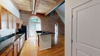 This two-floor loft-style space masterfully combines historic industrial character with contemporary design. The main floor serves as the heart of the home, where a spacious open-plan kitchen and living area commands immediate attention. The kitchen features a large central island with dark granite countertops, warm wood cabinetry, stainless steel appliances, and track lighting beneath exposed wood-beam ceilings with visible ductwork — a hallmark of its industrial aesthetic. Striking arched brick windows flood the space with natural light and frame lush outdoor greenery, while original red brick walls add rich texture and warmth throughout. Hardwood floors extend across the main level, tying the living and kitchen areas together seamlessly.

A staircase with modern metal railings leads to the upper floor, a carpeted loft-style area that offers versatility as a bedroom or additional living space. This level continues the brick wall detailing and features its own arched window, along with a bank of large modern windows overlooking the treetops. The upper floor also provides access to the full bathroom, which is finished with tile flooring, a tub-shower combination with white subway tile surround, and warm wood vanity cabinetry. Together, the two floors create a distinctive and characterful living environment.