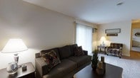 This single-floor space centers around a comfortable and inviting living area designed with a warm, neutral color palette throughout. The living room is anchored by a substantial dark upholstered sofa dressed with colorful geometric-patterned throw pillows, adding a touch of personality to the understated décor. A large window dressed with vertical blinds allows natural light to filter softly into the room, creating a bright and airy atmosphere. A wall-mounted air conditioning unit is positioned conveniently above the window, ensuring year-round comfort. The flooring transitions from dark hardwood in the main seating area to carpet in an adjoining nook or sitting area, which features a classic side table and lamp that cast a warm, ambient glow. Decorative elements, including a potted plant and sculptural accents on the coffee table, bring life and texture to the space. The clean, smooth ceiling and well-maintained walls contribute to an overall sense of tidiness and simplicity. The layout is efficient and functional, making excellent use of the available space while maintaining an open and uncluttered feel throughout.
