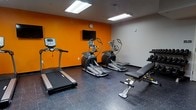 This fitness room is designed to support a well-rounded exercise routine within a compact, efficiently organized space. The room features a bold orange accent wall that adds energy to the environment, contrasting with the neutral gray walls and dark rubber flooring, which provides durability and traction underfoot. Overhead, flush-mounted fluorescent light fixtures ensure the space is well-lit throughout.

The cardio equipment lineup includes a treadmill positioned along one wall and two elliptical cross-trainers arranged side by side, offering users a variety of low-impact and higher-intensity options. A wall-mounted flat-screen television is positioned at a comfortable viewing angle, allowing users to stay entertained during workouts. An adjustable weight bench is situated toward the front of the room, accompanied by a rack of dumbbells, catering to strength training needs.

The overall layout is practical and straightforward, maximizing the available floor space while keeping equipment accessible. The single-floor setup ensures easy entry and exit, making this fitness room a convenient option for those seeking a no-frills workout environment.