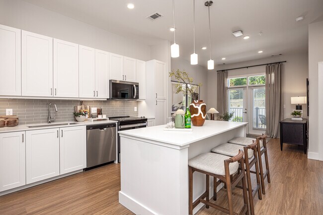 Cocina moderna con gabinetes blancos, iluminación bajo los gabinetes, protector contra salpicaduras de azulejos grises y encimeras de cuarzo blanco. - Avalon Carmel Commons