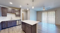 This single-floor unit offers a thoughtfully designed open layout that seamlessly connects the kitchen and living areas. The kitchen is a standout feature, showcasing rich dark-toned shaker-style cabinetry, crisp white quartz countertops, and a full suite of stainless steel appliances including a refrigerator, range, microwave, and dishwasher. A subway tile backsplash adds a clean, contemporary touch, while a central island with pendant lighting above provides additional prep space and a natural gathering point. Light wood-style flooring runs throughout the main living areas, complementing the neutral wall tones and creating a warm, cohesive aesthetic. The living area is bright and airy, enhanced by a ceiling fan and a sliding glass door with vertical blinds that opens to an outdoor space, inviting natural light throughout the day. The bedrooms feature soft carpeting, ceiling fans, and windows fitted with blinds, offering a comfortable and restful environment. Multiple doorways within the bedroom areas suggest closet storage and ensuite or adjacent bathroom access. Throughout the unit, clean lines, recessed lighting, and a consistent neutral palette contribute to a modern, move-in-ready atmosphere suited for everyday living.