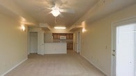 This single-floor space features an open-concept layout that seamlessly connects the living area to the kitchen, creating a functional and cohesive environment. The living area is finished with plush wall-to-wall carpeting in a neutral tone, contributing to a warm and understated ambiance. A ceiling fan with an integrated light fixture is centrally mounted, providing both air circulation and overhead lighting.

The kitchen is positioned at the rear of the open space and is defined by a raised breakfast bar that serves as a natural divider between the cooking and living zones. Warm wood-toned cabinetry lines the kitchen walls, complemented by countertops and standard white appliances, including a built-in microwave. Recessed fluorescent lighting above the kitchen area ensures ample task illumination.

Multiple doorways are visible throughout the space, indicating access to additional rooms and storage areas, including what appears to be a spacious walk-in closet. The walls throughout are finished in a soft, neutral tone, and the overall design prioritizes practicality and ease of movement. The layout is well-suited for everyday living, offering a clean and straightforward aesthetic.