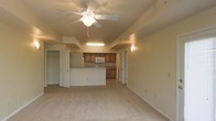 This single-floor space features an open-concept layout that seamlessly connects the living area to the kitchen, creating a functional and cohesive environment. The living area is finished with plush wall-to-wall carpeting in a neutral tone, contributing to a warm and understated ambiance. A ceiling fan with an integrated light fixture is centrally mounted, providing both air circulation and overhead lighting.

The kitchen is positioned at the rear of the open space and is defined by a raised breakfast bar that serves as a natural divider between the cooking and living zones. Warm wood-toned cabinetry lines the kitchen walls, complemented by countertops and standard white appliances, including a built-in microwave. Recessed fluorescent lighting above the kitchen area ensures ample task illumination.

Multiple doorways are visible throughout the space, indicating access to additional rooms and storage areas, including what appears to be a spacious walk-in closet. The walls throughout are finished in a soft, neutral tone, and the overall design prioritizes practicality and ease of movement. The layout is well-suited for everyday living, offering a clean and straightforward aesthetic.