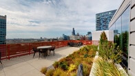 Located atop 51 S 14th Street in Chicago's South Loop, this rooftop deck offers an expansive open-air retreat with commanding, panoramic views of the Chicago skyline. The Willis Tower and surrounding high-rises create a dramatic urban backdrop visible from nearly every vantage point on the deck.

The rooftop is defined by a generous concrete paver terrace that provides ample space for outdoor gathering and relaxation. Multiple seating arrangements are distributed across the deck, including round bistro tables with chairs and low-profile lounge furniture, allowing for both casual conversation and more formal outdoor dining.

A standout feature of this rooftop is its integrated green roof planting system. Raised concrete planters and ground-level green beds are filled with ornamental grasses, low shrubs, succulents, and mixed perennials, adding texture, color, and a naturalistic quality to the urban setting. The plantings are thoughtfully arranged to complement the open paved areas without obstructing the skyline sightlines.

A bold red perimeter railing runs along the edges of the deck, providing safety while contributing a strong visual design element. Floor-to-ceiling glass doors offer seamless access to the interior, and the glazed facade reflects the surrounding cityscape. The deck enjoys abundant natural light and open sky exposure, making it a striking and functional amenity space in the heart of Chicago.