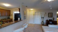 This single-story home on Hearthside Dr in Wilmington, NC, presents a comfortable and functional layout with a warm, neutral color palette throughout. The living area is finished with plush carpeting and benefits from ambient ceiling lighting, creating an inviting atmosphere. A partial wall with a tiled breakfast bar separates the living space from the kitchen, which features dark cabinetry, a refrigerator, and a window with horizontal blinds that allows natural light to filter in. Decorative touches, including a tall vase with bamboo stems and a framed mirror, add character to the space. Multiple interior doors—finished in classic six-panel white—provide access to additional rooms and closets, contributing to a well-organized floor plan. The entryway area is accented with a wall-mounted key and accessory rack, offering practical storage near the entrance. White modern bar stools complement the breakfast bar, and a portion of a white sofa is visible, suggesting a generously sized living area. Overall, the home offers a clean, move-in-ready aesthetic suited to everyday living in coastal North Carolina.