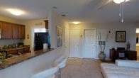 This single-story home on Hearthside Dr in Wilmington, NC, presents a comfortable and functional layout with a warm, neutral color palette throughout. The living area is finished with plush carpeting and benefits from ambient ceiling lighting, creating an inviting atmosphere. A partial wall with a tiled breakfast bar separates the living space from the kitchen, which features dark cabinetry, a refrigerator, and a window with horizontal blinds that allows natural light to filter in. Decorative touches, including a tall vase with bamboo stems and a framed mirror, add character to the space. Multiple interior doors—finished in classic six-panel white—provide access to additional rooms and closets, contributing to a well-organized floor plan. The entryway area is accented with a wall-mounted key and accessory rack, offering practical storage near the entrance. White modern bar stools complement the breakfast bar, and a portion of a white sofa is visible, suggesting a generously sized living area. Overall, the home offers a clean, move-in-ready aesthetic suited to everyday living in coastal North Carolina.