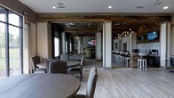 This single-floor communal space blends rustic charm with modern sophistication in a seamlessly connected open-plan layout. Reclaimed wood ceiling beams and paneled accents set a warm, inviting tone throughout, complemented by light wide-plank hardwood flooring that brightens the interior. The main gathering area features generous round dining tables paired with upholstered side chairs in neutral gray tones, creating an approachable and comfortable setting for socializing. Recessed lighting and stylish geometric pendant fixtures provide layered illumination across the space. Moving deeper into the layout, a billiards area offers a dedicated recreation zone, anchored by a full-size pool table and a wall-mounted television, making it ideal for casual entertainment. Adjacent to this, a dramatic bar counter stretches along one wall, lined with evenly spaced bar stools beneath a canopy of Edison-style pendant lights that lend a warm, ambient glow. Mirrors along the bar wall enhance the sense of depth and openness. Structural white columns define zones without closing off sightlines, maintaining an airy, flowing feel throughout. The overall ambiance is one of relaxed elegance — a well-appointed space designed for both lively gatherings and laid-back leisure.