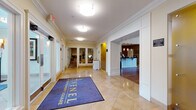 Avenel at Montgomery Square is a well-appointed residential community offering a thoughtfully designed amenity floor that caters to both comfort and productivity. The main entrance greets residents and guests with a spacious lobby featuring elegant travertine tile flooring, crown molding, and ample overhead lighting that creates a bright, polished first impression. A branded entry mat anchors the corridor, which connects seamlessly to the community's key amenity spaces.

Adjacent to the lobby is a stylish lounge and café station, where a white Shaker-style counter with a dark granite top is complemented by pendant lighting and warm wood floors. A striking stone fireplace with a rustic wood mantel serves as the centerpiece of the common lounge area, adding a cozy, inviting ambiance enhanced by hardwood flooring and large windows that bring in natural light.

The fitness center is generously equipped with cardio machines including treadmills and ellipticals, a cable weight station, free weights, and adjustable benches, all set on durable rubber flooring with mirrored walls to maximize the sense of space.

The business center offers multiple computer workstations along a shared counter, comfortable task chairs, and access to a multifunction copier/printer, making it a practical resource for remote work or administrative needs. A smaller breakout room with a round table and large windows provides a quiet space for meetings or focused work.