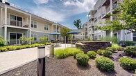 Located at 715 US Highway 46, this outdoor courtyard space is a well-designed communal area nestled within a multi-story residential building complex. The courtyard is thoughtfully landscaped, featuring expansive stretches of vibrant green lawn framed by carefully maintained garden beds filled with ornamental grasses, flowering shrubs, boxwood hedges, and seasonal plantings. Stone-clad raised planters and retaining walls add architectural texture and visual interest throughout the space.

A central patio area provides an inviting gathering spot, furnished with outdoor tables, chairs, and shade umbrellas, ideal for relaxation or socializing. Curved and straight pathways of light-toned pavement wind through the greenery, connecting various areas of the courtyard. Benches are thoughtfully placed to offer quiet seating with views of the surrounding landscape.

The surrounding multi-story building, clad in a combination of light beige and gray horizontal siding, features large windows, private balconies, and covered ground-floor entries that open directly to the courtyard. The architecture creates an enclosed, sheltered feel while maintaining an open-air environment filled with natural light. The overall ambiance is serene and resort-like, offering residents a welcoming outdoor retreat with a strong sense of community and connection to nature.