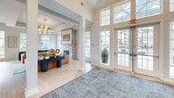 Located at 18237 Tempo Boulevard in Westfield, Indiana, this single-story community amenity facility offers an expansive and thoughtfully designed layout across a generously sized floor plan. The living room serves as the heart of the space, boasting soaring ceilings, large multi-pane windows that flood the area with natural light, and views of the surrounding outdoor environment. A sleek tile-surround fireplace anchors one wall, while contemporary gold-toned chandeliers add a refined elegance. Comfortable seating areas and decorative accents create an inviting, upscale atmosphere. Adjacent to the living area, a modern kitchen features a long quartz waterfall island with bar seating, white shaker cabinetry, stainless steel appliances, and classic subway tile backsplash. The hallway connects the various zones of the facility seamlessly, lined with additional lounge seating and workstation booths beneath recessed lighting and accent walls in deep green. The two exercise rooms are generously sized and well-appointed — one outfitted with treadmills, ellipticals, rowing machines, and cable equipment on durable rubberized flooring with soft teal walls and outdoor views, and the other serving as a studio-style space with mirrored walls, a wall-mounted TV, cycling equipment, a heavy bag, and a ceiling fan overhead.