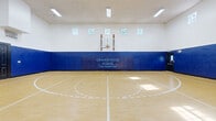 The indoor sports court at Grandewood Pointe Luxury Apartment Homes, located in Orlando, Florida, is a generously sized, full-scale basketball facility designed to accommodate serious athletic activity. The court features a polished hardwood-style floor with crisp, clearly marked regulation lines, including a center circle, three-point arcs, and free-throw lanes, providing a professional playing surface for residents.

Multiple glass-backboard basketball hoops are mounted along opposing ends and sides of the court, allowing for versatile gameplay configurations. The walls are lined with thick, blue padded safety panels that extend to mid-wall height, bearing the Grandewood Pointe branding — a practical and aesthetic design choice that reinforces both player safety and community identity.

Soaring ceilings elevate the sense of openness within the space, while a row of high clerestory windows along the upper walls admits natural light, supplemented by banks of overhead fluorescent lighting to ensure consistent illumination during all hours of use. A posted sports court rules sign near the entrance helps maintain an organized and respectful environment.

An adjacent mirrored fitness area is visible through a doorway, indicating that the sports court is part of a larger amenity complex. The space is well-maintained and thoughtfully designed, reflecting the community's commitment to offering premium recreational facilities.