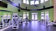 This fitness center is housed within a striking single-floor space defined by its dramatic double-height ceilings and an abundance of natural light. The room's bold lime green walls are accented by a dark horizontal band that runs the perimeter, adding a dynamic visual element to the interior. A large ceiling fan is centrally mounted overhead, promoting air circulation throughout the workout area. Rows of upper-level clerestory windows, paired with full-sized lower windows and elegant double glass-paned entry doors, flood the space with natural light and offer views of the surrounding landscape. The durable rubber flooring is well-suited for fitness use, providing traction and cushioning across the generous open floor plan. The facility is outfitted with a variety of strength training machines and cardio equipment, including weight machines, an elliptical trainer, and a rowing machine, offering users a comprehensive workout experience. Wall-mounted mirrors along one side enhance the sense of space and allow for form monitoring during exercise. Mounted televisions are also present, adding to the functional amenities of the facility. The overall atmosphere is energetic yet organized, making it a practical and inviting environment for fitness enthusiasts.