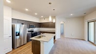Located at 9915 Dessau Rd in Austin, TX, this thoughtfully designed one-bedroom, one-bathroom residence offers a comfortable and functional single-floor layout. The open-concept living and kitchen area creates a seamless flow, anchored by a kitchen featuring rich dark-toned cabinetry, granite countertops, a subway tile backsplash, and a full suite of stainless steel appliances including a refrigerator, range, and microwave. A peninsula with a built-in sink and pendant lighting adds both style and practicality, while wood-look flooring grounds the kitchen space. The living area is carpeted and bathed in natural light, with a sliding glass door providing outdoor access and views of the surroundings. The bedroom is a well-sized, carpeted retreat with a ceiling fan, a window with natural light, and a sliding door. It connects to a walk-in closet outfitted with wire shelving and a central shelving tower for organized storage. The bathroom features a granite-topped vanity with dark cabinetry, a tub-shower combination, and wood-look flooring. A dedicated laundry area with a full-size washer and overhead wire shelving adds everyday convenience. Neutral tones and recessed lighting throughout contribute to a clean, modern ambiance.