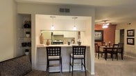 This single-floor residence is thoughtfully designed to balance functionality and comfort. The kitchen serves as a central highlight, featuring white cabinetry that extends to the ceiling for ample storage, and a built-in range with an overhead hood. A peninsula-style breakfast bar with a light countertop extends outward, accommodating two bar-height chairs with cushioned seats — ideal for casual dining or conversation. Two pendant lights hang above the bar, providing focused task lighting that adds a warm, inviting glow to the space. Overhead, a ceiling vent ensures efficient climate control throughout. Adjacent to the kitchen, the dining area is distinguished by a rich terracotta accent wall that creates a cozy, intimate atmosphere. A ceiling fan with a light fixture services this area, while framed artwork and a formal dining set complete the look. The flooring transitions from carpet in the living areas to the kitchen zone, adding texture and definition between spaces. The overall layout is open and connected, allowing for easy flow between the kitchen, dining, and living areas, making this residence well-suited for both everyday living and entertaining.