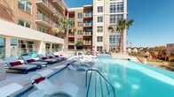 Located at 1303 Montrose Blvd in Houston's vibrant Montrose neighborhood, this outdoor amenity deck offers a resort-inspired experience in the heart of the city. The centerpiece of the space is a large, shimmering swimming pool with a shallow wading shelf lined with sleek white in-pool loungers accented by red cushions, ideal for relaxing in the Texas sun. The expansive pool deck is surrounded by additional lounge chairs and chaise lounges, providing ample space for sunbathing and socializing. Tall, tropical palm trees are strategically placed throughout the deck, lending a warm, resort-like atmosphere. The pool area features glass railing along one edge, offering unobstructed views of the Houston skyline and the surrounding Montrose and Midtown neighborhoods. A separate covered patio area adjacent to the pool provides a shaded retreat, equipped with comfortable outdoor seating, dining furniture, mounted outdoor televisions, and string lighting overhead, creating an inviting environment for both daytime lounging and evening gatherings. The building's contemporary brick and stucco façade rises impressively above the deck, with private balconies overlooking the amenity space. The entire outdoor area is finished with large-format light-toned tile pavers, contributing to a clean, upscale aesthetic throughout.