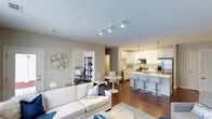 This single-floor residence showcases a well-designed open-concept layout that connects the living, dining, and kitchen areas into one cohesive, flowing space. The living room is anchored by a plush white sofa accented with blue and white throw pillows, complemented by a soft area rug and decorative wall art, creating a comfortable and stylish atmosphere. Track lighting runs across the ceiling, providing consistent illumination throughout the main living area.

The kitchen is a standout feature, outfitted with white shaker-style cabinetry, stainless steel appliances including a refrigerator, range, and microwave, and a peninsula breakfast bar lined with modern counter-height stools. Pendant lighting above the bar adds a refined touch. A compact dining area with a round white table and sleek white chairs sits adjacent to the kitchen, ideal for casual meals.

A doorway visible from the living room leads to an additional room, partially revealed as a home office or bedroom with dark shelving and a desk setup. The warm hardwood flooring ties the entire space together, contributing to the overall clean, modern, and inviting ambiance.