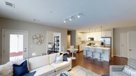 This single-floor residence showcases a well-designed open-concept layout that connects the living, dining, and kitchen areas into one cohesive, flowing space. The living room is anchored by a plush white sofa accented with blue and white throw pillows, complemented by a soft area rug and decorative wall art, creating a comfortable and stylish atmosphere. Track lighting runs across the ceiling, providing consistent illumination throughout the main living area.

The kitchen is a standout feature, outfitted with white shaker-style cabinetry, stainless steel appliances including a refrigerator, range, and microwave, and a peninsula breakfast bar lined with modern counter-height stools. Pendant lighting above the bar adds a refined touch. A compact dining area with a round white table and sleek white chairs sits adjacent to the kitchen, ideal for casual meals.

A doorway visible from the living room leads to an additional room, partially revealed as a home office or bedroom with dark shelving and a desk setup. The warm hardwood flooring ties the entire space together, contributing to the overall clean, modern, and inviting ambiance.