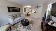 This single-floor residence offers a thoughtfully designed open-concept layout that seamlessly connects the main living areas. The living room is a welcoming, comfortably scaled space featuring plush wall-to-wall carpeting, neutral warm-toned walls, and ample room for relaxed seating arrangements. Decorative wall art and classic crown molding add refined touches to the space.

The living area flows naturally into a dedicated dining space, which is defined by elegant wainscoting along the lower walls and anchored by a stylish pendant light fixture overhead. The dining area comfortably accommodates a full table and chair set, making it well-suited for both everyday meals and entertaining.

Adjacent to the dining space, a pass-through kitchen connects the cooking area to the rest of the home, providing a practical and interactive layout. The kitchen features light-toned cabinetry and countertops, contributing to a bright and functional workspace.

Throughout the main living areas, the neutral color palette and consistent flooring create a cohesive, inviting atmosphere. The open sightlines between rooms enhance the sense of space and allow natural light to travel freely, making the entire floor feel airy and well-connected. The overall design balances functionality with a clean, classic aesthetic.