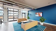 This contemporary single-floor space blends industrial design with modern comfort, creating an inviting and functional amenity lounge. The open-plan layout is anchored by a spacious seating area furnished with tan leather sofas and a teal geometric area rug, complemented by a bold cobalt blue accent wall hosting a wall-mounted flat-screen display. Exposed metallic ductwork runs overhead, lending an industrial character to the otherwise clean, modern aesthetic.

Natural light floods the space through large floor-to-ceiling glass garage-style doors that open onto an outdoor terrace, offering expansive urban skyline views. Additional clerestory windows line the walls, ensuring consistent brightness throughout. The secondary seating zone features plush cream accent chairs, globe and arc floor lamps, and circular wall sconces, creating a more intimate atmosphere within the broader open space.

The well-appointed kitchen area is finished with light wood cabinetry, warm butcher block countertops, and a striking blue backsplash that ties cohesively to the lounge's color palette. Open shelving, track lighting, stainless steel appliances, and ample counter space make it highly functional. Rich wood-look flooring flows continuously throughout, tying all areas together seamlessly. Lush potted plants are strategically placed, adding a refreshing natural element to the space's contemporary urban design.
