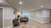 This single-floor common area is designed to serve as a functional and comfortable shared space for residents. The lobby lounge is the centerpiece of the area, furnished with a tufted gray sofa, coordinating armchairs, and a wire-frame accent table, creating an inviting seating arrangement for residents to relax or gather. The space is finished with wood-look porcelain tile flooring that runs throughout, lending warmth and a cohesive aesthetic to the entire floor.

Neutral gray walls are accented by white chair rail molding and crisp white baseboards, contributing to a clean and polished appearance. A drop tile ceiling with recessed can lighting provides consistent, even illumination across the space. Decorative touches such as a wall clock, a round mirror, and potted greenery add character without overwhelming the functional nature of the area.

A bank of individual mailboxes lines one wall, offering a convenient and organized mail retrieval area for residents. The stairwell, framed by white-painted wood railings with a dark handrail, connects to additional levels and is positioned near a set of glass-panel double doors that allow natural light to filter into the corridor. The hallway leads to multiple doorways, providing access to additional rooms or units. Overall, the space balances practicality with a refined, residential aesthetic.