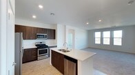 This single-floor space showcases a thoughtfully designed open-concept layout that seamlessly connects the kitchen and living areas. The kitchen is a standout feature, outfitted with rich, dark espresso-toned cabinetry that provides a striking contrast against the crisp white countertops. A center island with a double stainless steel sink serves as both a functional workspace and a natural gathering point. Stainless steel appliances, including a gas range with oven, over-the-range microwave, and dishwasher, complete the kitchen's modern and efficient setup. Recessed lighting throughout the ceiling provides consistent, bright illumination across the space. Light-toned flooring in the kitchen transitions into carpeting in the adjacent living area, subtly defining each zone while maintaining the open feel. The living area benefits from a large window that invites natural light deep into the space, enhancing the clean, airy ambiance. Neutral white walls throughout keep the interiors feeling fresh and versatile. A doorway visible from the main living area hints at additional rooms beyond, rounding out a well-connected and functional floor plan.