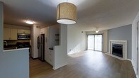 This single-floor residence offers a thoughtfully designed open-concept layout that seamlessly connects the kitchen and living areas. The kitchen is outfitted with tall white cabinetry, open display shelving, and a stainless steel refrigerator, providing ample storage and a clean, modern aesthetic. A stylish drum pendant light hangs overhead, adding a warm focal point to the space. A half-wall partition subtly defines the kitchen from the rest of the living area while maintaining an open and airy feel throughout. The living area is generously proportioned, featuring smooth hardwood-style flooring that extends throughout the unit, contributing to a cohesive and contemporary look. A ceiling fan with a light fixture keeps the space comfortable, while a full-length sliding glass door floods the room with natural light and offers potential outdoor access. Vertical blinds provide privacy and light control. The textured ceiling adds a classic touch, and neutral wall tones create a versatile backdrop suitable for a variety of interior styles. The overall ambiance is bright, clean, and welcoming, making this a functional and attractive single-level living space.