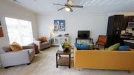 This well-appointed single-floor space offers a warm and inviting atmosphere anchored by a thoughtfully designed living room. The room features classic crown molding that frames the ceiling, adding an elegant architectural touch, while a ceiling fan with integrated lighting provides both comfort and functionality. Soft, neutral carpeting runs throughout, lending a cozy feel underfoot.

The living area is furnished with a bold mustard-yellow sofa accented by vibrant teal and green throw pillows, complemented by a pair of grey accent chairs and a rich wooden coffee table with a lower shelf for added storage. A white lattice-door sideboard serves as a stylish media console, topped with decorative accents and a flat-screen television. An additional accent chair in warm orange tones adds a pop of color to the corner.

Natural light filters in through a window, brightening the space and highlighting the cheerful, eclectic decor — including a vivid blue artwork depicting cows, which serves as a striking focal point on the wall. The overall ambiance is lively yet comfortable, blending classic architectural details with a colorful, contemporary furnishing palette that creates an energetic and welcoming environment.