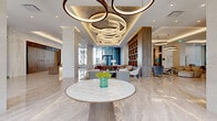 The Carlyle's lobby at 815 Avenue Plymouth in Mont-Royal, Quebec, makes an immediate impression with its sophisticated, hotel-caliber design. Polished large-format marble flooring stretches throughout the expansive space, reflecting abundant natural light that pours in through floor-to-ceiling glazed façades offering views of the surrounding streetscape and greenery. At the heart of the entrance, a sculptural multi-ring chandelier suspended from a coffered ceiling serves as a striking focal point, casting warm ambient light across the space. A sleek concierge desk, clad in warm wood tones and bearing the Carlyle branding, provides a welcoming point of arrival. The lobby flows seamlessly into several distinct lounge and seating areas furnished with contemporary neutral-toned sofas, accent chairs, and refined coffee tables. A dramatic floor-to-ceiling built-in shelving wall in rich walnut wood anchors one side of the space, adding warmth and visual depth. Curated artwork and tasteful floral arrangements contribute to the refined atmosphere throughout. The overall design palette of warm beiges, soft whites, and natural wood tones creates a cohesive, understated elegance, establishing the Carlyle lobby as a welcoming and luxurious communal space.