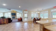 Located in Wales Crossing, North Canton, Ohio, this single-floor community clubhouse offers a welcoming and well-appointed gathering space designed for comfort and functionality. The centerpiece of the space is a spacious great room anchored by a striking floor-to-ceiling stone fireplace with a wall-mounted television above, soaring vaulted ceilings, and a ceiling fan — creating a warm, inviting atmosphere. Multiple seating arrangements, including leather sofas and armchairs, are positioned throughout the lounge areas, which are bathed in natural light streaming through an abundance of large windows with transom panels. The open layout seamlessly connects the lounge to a dedicated dining area, complete with a wood dining table, upholstered chairs, and an elegant chandelier overhead. Adjacent to the dining space is a fully equipped kitchen featuring warm honey-toned cabinetry, light stone countertops, stainless steel appliances including a dishwasher, double-drawer warming unit, built-in microwave, and a ceiling fan — all set atop neutral tile flooring that extends throughout the common areas. Crown molding and soft yellow walls lend a cohesive, refined aesthetic across the entire space, while exterior views of lush greenery are visible from nearly every room, reinforcing the community's serene residential setting.