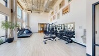 Located in South Salt Lake, Utah, this well-appointed fitness center offers an expansive, single-floor exercise space designed to support a full range of workout routines. The room is defined by its dramatic double-height ceilings with an exposed industrial-style ceiling structure, lending the space an open, airy feel that is further amplified by an abundance of natural light.

A striking wall of floor-to-ceiling windows dominates one side of the gym, framing sweeping views of the surrounding landscape and the Wasatch Mountain Range beyond—providing an inspiring backdrop during any workout. Light hardwood-style flooring runs throughout, contributing to the clean, modern aesthetic.

The cardio section is well-equipped with multiple treadmills, a stair climber, elliptical trainers, stationary bikes, and rowing machines, all positioned to take advantage of the panoramic views. The strength training area features a multi-station cable machine, adjustable weight benches, and a comprehensive dumbbell rack. A built-in water fountain station provides added convenience, while framed motivational artwork adorns the walls, adding character to the space. A separate adjacent room visible through glass doors offers additional utility. The overall ambiance is bright, energizing, and thoughtfully designed for an optimal fitness experience.