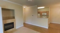 This single-story space showcases a practical and open floor plan that seamlessly connects the living and kitchen areas. The living space features a combination of hardwood flooring and carpet, accented by clean white baseboards and elegant crown molding that runs along the ceiling, adding a refined architectural detail throughout. A tile-surround fireplace anchors one side of the living area, providing both a functional and aesthetic focal point.

The kitchen is positioned adjacent to the living area and is accessible via a raised breakfast bar with a light-toned countertop, encouraging an open and social atmosphere. The kitchen is equipped with medium-toned wood cabinetry, a white refrigerator, a white microwave, and a gas range, offering practical functionality. A utility light fixture above the kitchen provides ample task lighting.

A flush-mount ceiling light fixture illuminates the living area, while neutral, warm-beige walls throughout create a cohesive and inviting ambiance. A hallway visible in the background leads to additional areas of the unit, suggesting a well-organized layout. Overall, the space presents a clean, move-in-ready environment with classic detailing.