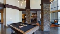 This single-floor amenity lounge makes a striking first impression with its soaring double-height ceilings and dramatic design language that blends rustic and contemporary elements throughout. Prominent stacked stone columns with rich, earthy tones anchor the space, lending a sense of structure and texture that flows seamlessly from one area to the next. A full-size billiards table with a dark felt surface serves as a centerpiece of the main recreation area, positioned beneath a cascading chandelier of exposed Edison-style bulbs that cast warm, ambient light across the room. Dark wood accents on the ceiling beams and column caps complement the natural stonework, creating a cohesive and inviting atmosphere. Beyond the columns, the space opens into a bar and lounge area, where additional pendant lighting and a backsplash of layered stone tiles continue the design motif. Bar seating and accent chairs in cool tones provide comfortable gathering spots for socializing. The open floor plan encourages a fluid transition between the game area and lounge, making the space well-suited for both casual recreation and social events. The overall ambiance is warm, upscale, and welcoming, with thoughtful material choices that give the space a refined yet relaxed character.